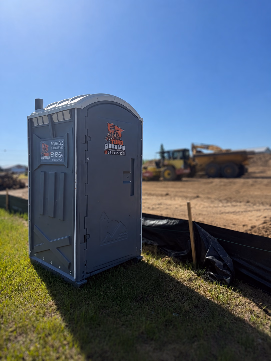 A gray portable toilet on a dirt construction site with earth-moving equipment behind it.