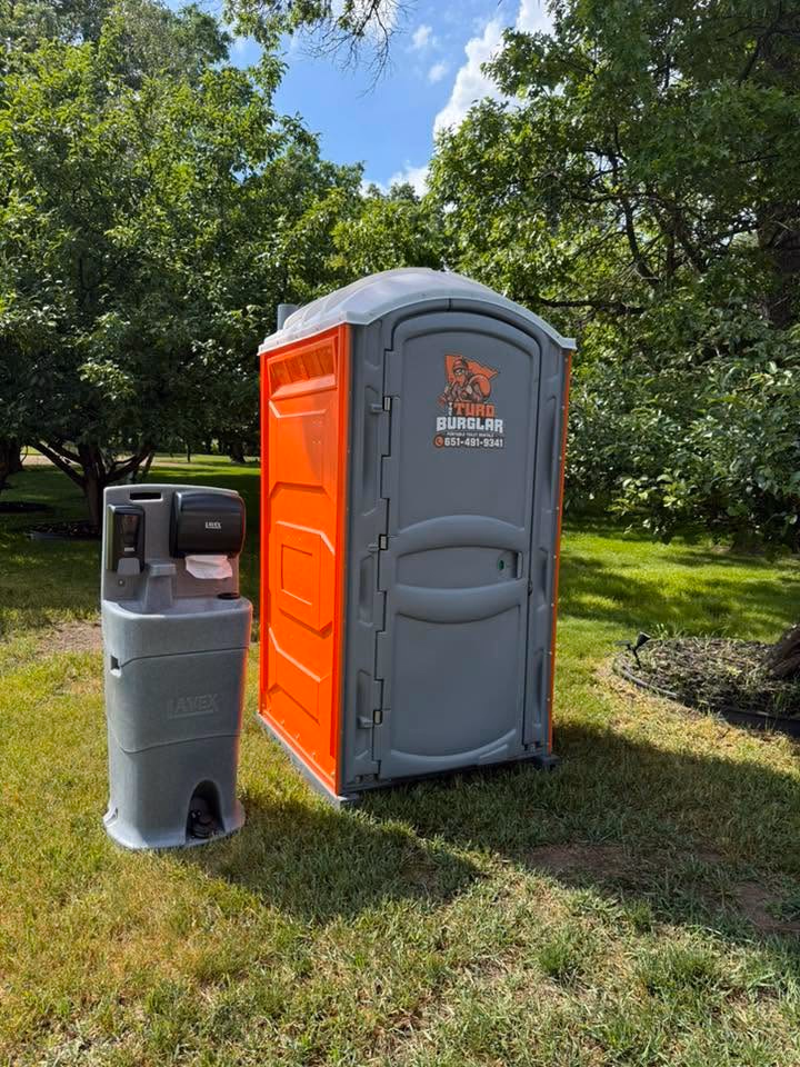 An orange and gray portable toilet beside a freestanding hand wash station on a grass lawn.