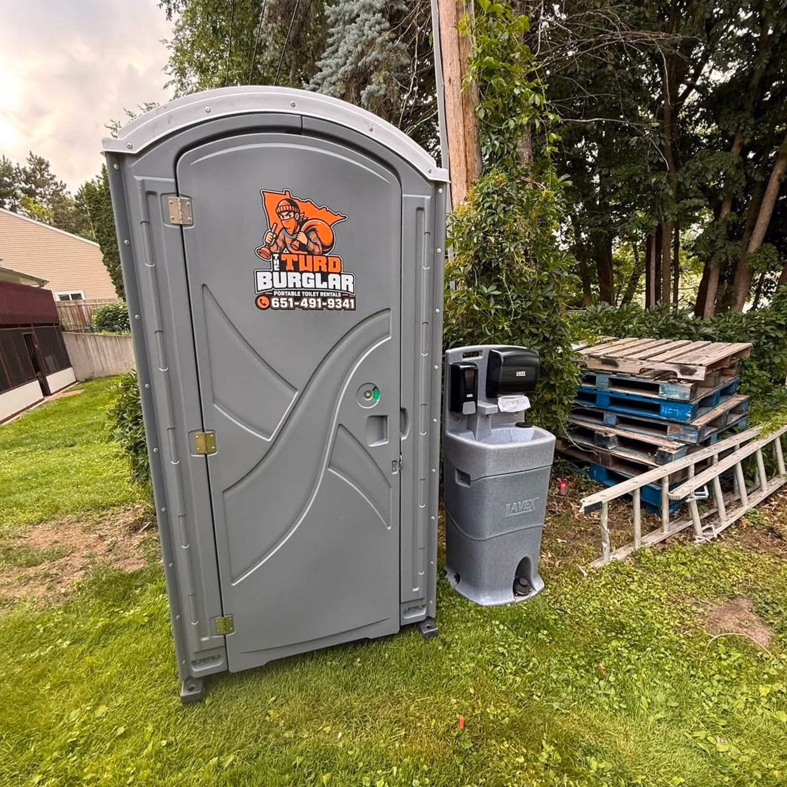 A gray portable toilet and a hand wash station set on grass beside a wooden ramp.