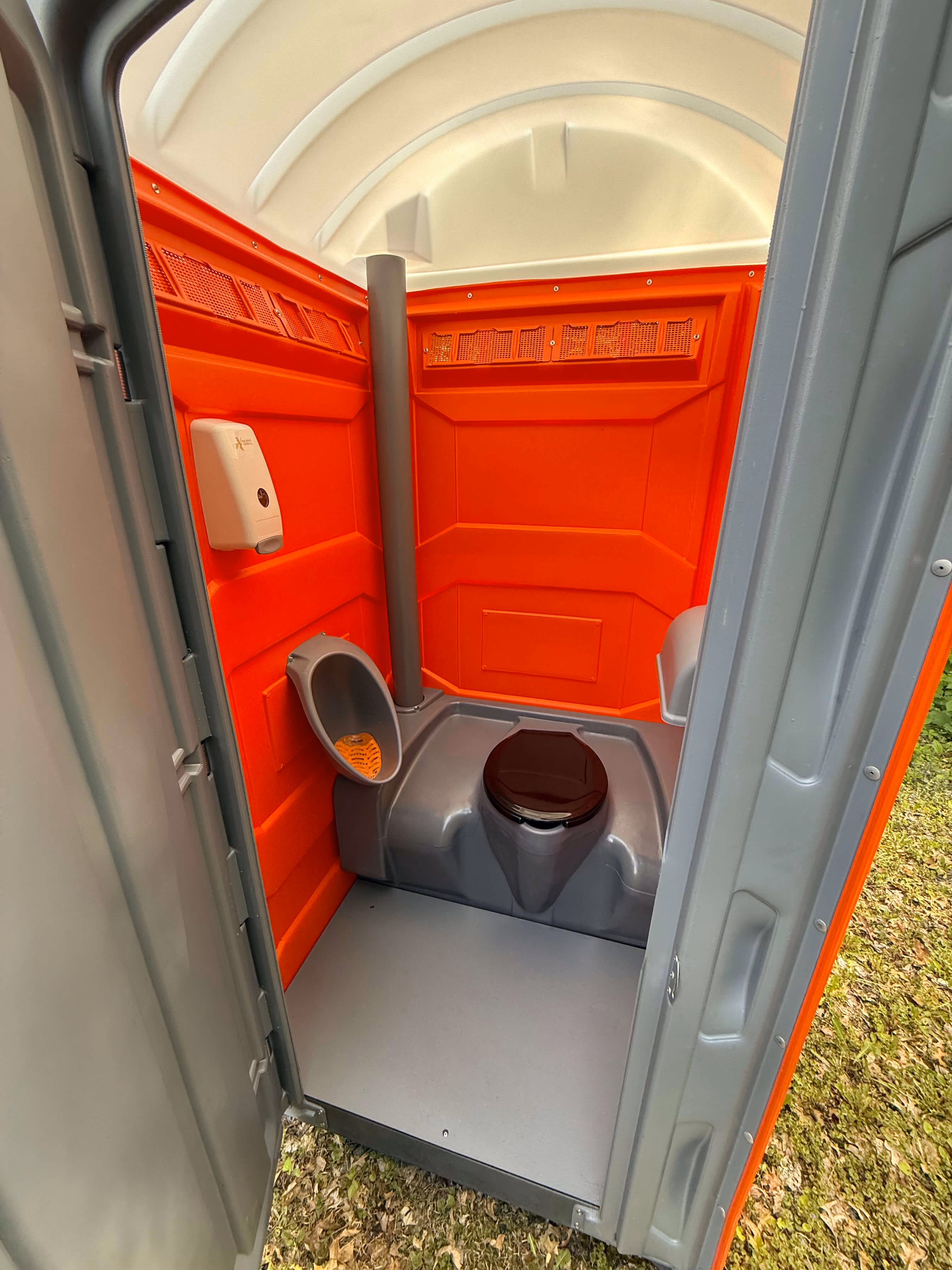 Inside a portable toilet, showing the orange walls, a urinal and a hand sanitizer dispenser with the door open to the grass.