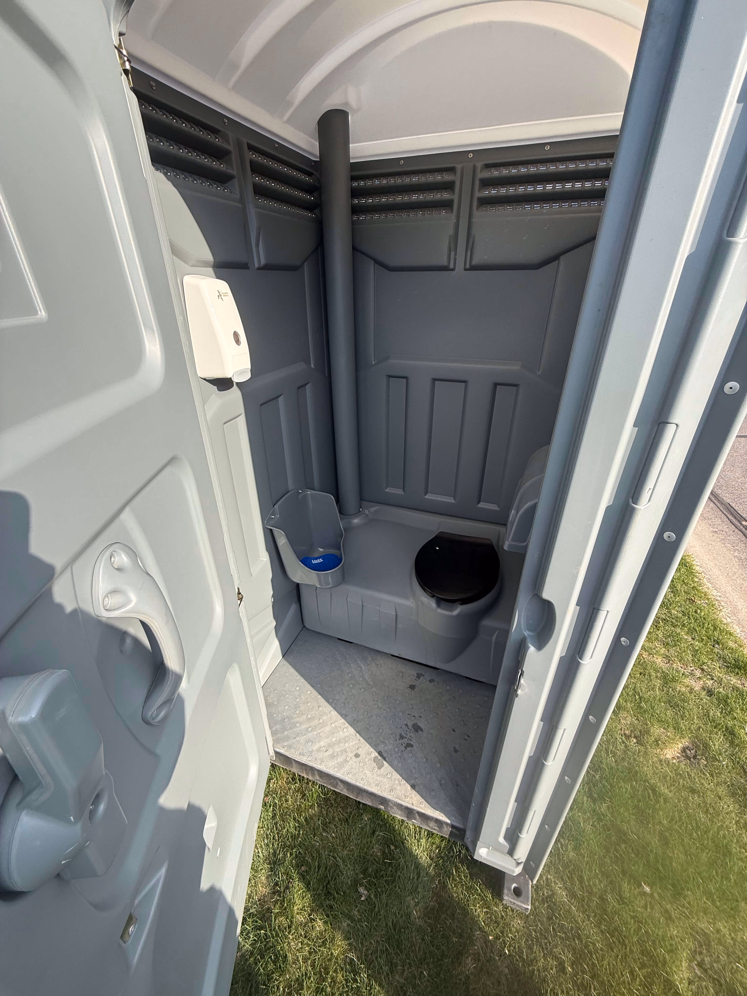 Inside a gray portable toilet with a wide open floor and a hand sanitizer dispenser beside the door.