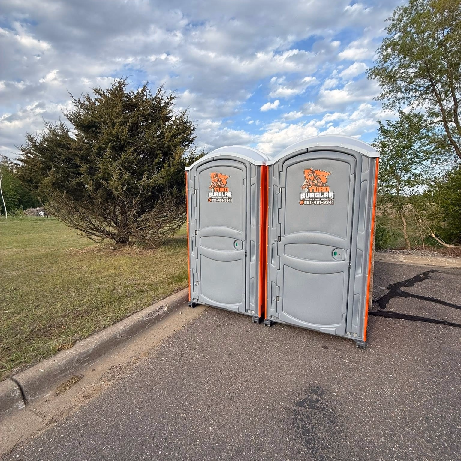 Alley beside a building with a gray portable toilet labeled Turd Burglar and a white service truck with coiled yellow and black hoses.