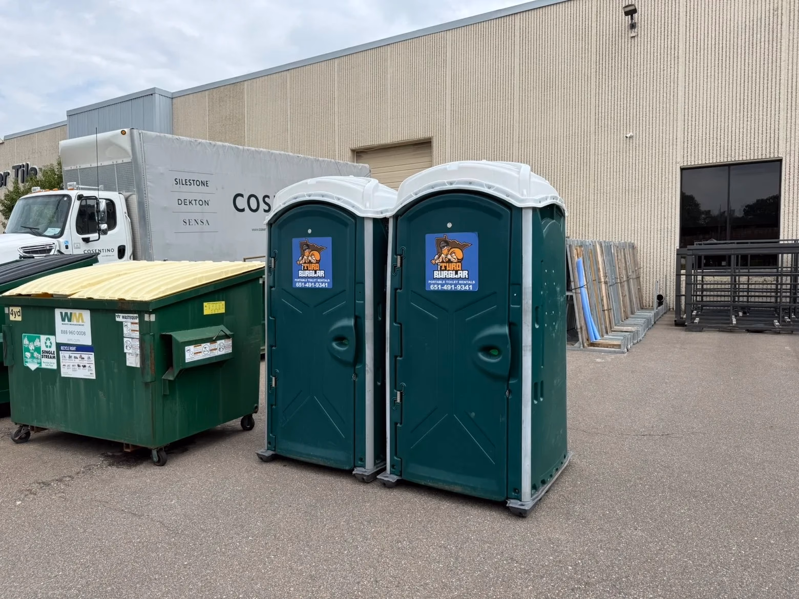 Two green portable toilets labeled 'Turd Burglar Portable Toilet Rentals' next to a green and yellow dumpster and a white truck parked by a beige warehouse building.